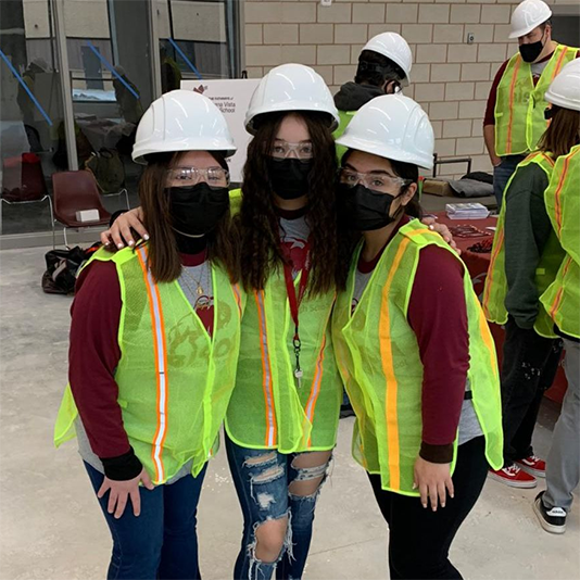  A group of young women wearing hardhats, high visibility vests, goggles and face coverings